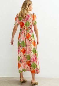 Kvinna klädd i en färgglad patchwork midi-klänning med korta puffärmar och gröna klacksandaler, stående mot en enkel vit vägg.