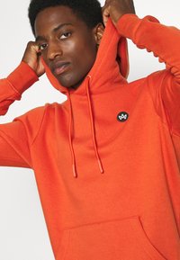 Młody mężczyzna regulujący kaptur jaskrawo pomarańczowej bluzy z małym czarno-białym logo na piersi.