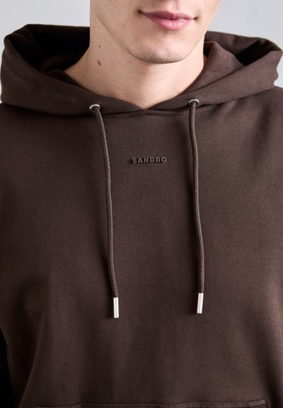 Felpa con cappuccio marrone realizzata in tessuto morbido, dotata di tasca frontale, chiusura con coulisse e punte in metallo, e un logo che recita "SANDRO."