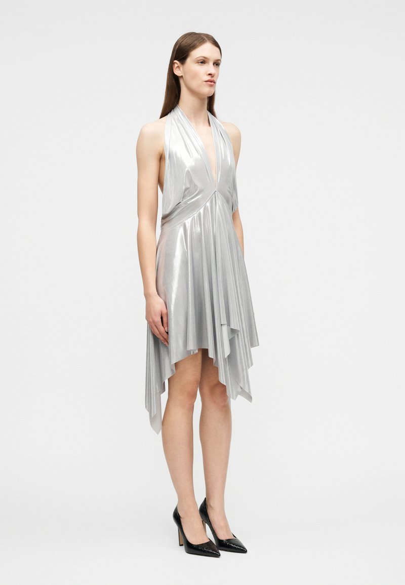 Femme portant une robe argentée sans manches à col halter avec un ourlet asymétrique et des escarpins noirs à bout pointu, se tenant devant un fond uni.