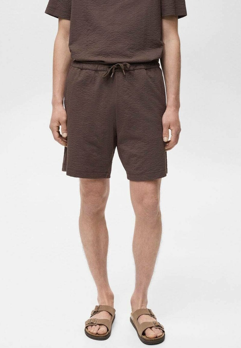 Shorts en tissu texturé marron avec une taille élastique et un cordon de serrage. Associés à un haut à manches courtes assorti et des sandales marron.