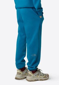 Tyrkysová mikina a zodpovedajúce tepláky z mäkkého materiálu, s jemným logom na nohavici a voľným strihom. Svetlosivé tenisky s sieťovinou.