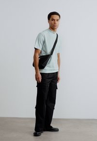 Γαλάζιο t-shirt με στρογγυλή λαιμόκοψη, μαύρη τσάντα χιαστί, μαύρο παντελόνι cargo και μαύρα παπούτσια πάνω σε απλό φόντο. Απλός, λειτουργικός σχεδιασμός.