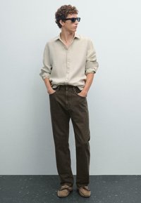 Casual outfit met een lichtbeige button-up shirt, donkerbruine jeans en tan suède schoenen met donkere veters. Zwarte zonnebril maakt de look compleet.