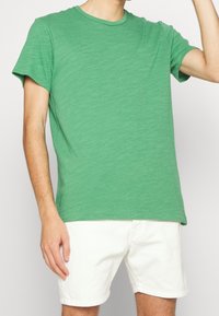 T-shirt vert à manches courtes en tissu doux et texturé avec un col rond ; porté avec un short blanc et tenu par une personne.
