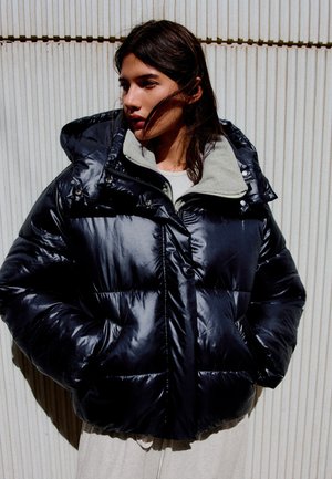 Femme portant une doudoune noire brillante avec capuche, debout contre un mur clair nervuré, regardant vers la gauche, les mains dans les poches.
