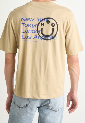 T-shirt en coton beige avec texte bleu listant des villes et un graphique de smiley noir dans le dos. Manches courtes, coupe décontractée, devant uni.