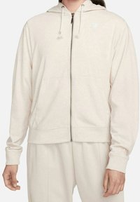 Ljust beige zip-up huvtröja i mjukt tyg, med dragsko på huvan, kängurufickor och en liten logotyp på vänster bröst.
