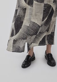 Robe longueur cheville à motifs noirs et blancs avec des boutons, présentant un ourlet fendu. Associée à des mocassins noirs brillants.