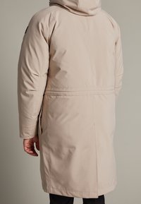 Beige vadderad jacka med huva, mittlång design, med sidofickor och textilstruktur. Sömdetaljer löper vertikalt längs ryggen.