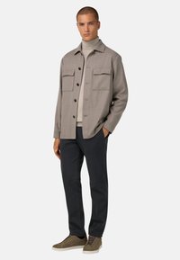 Šedá košile na knoflíky s dvěma náprsními kapsami, vrstvená přes béžový rolák. V kombinaci s tmavými kalhotami a olivově zelenými semišovými teniskami.