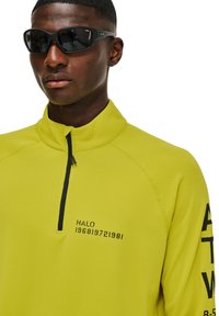 Jasnożółty pulower na ćwierć zamku z fakturowanej tkaniny, z czarnymi napisami na klatce piersiowej i prawym rękawie, oraz wysokim kołnierzem.