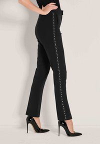 Zwarte op maat gemaakte broeken met een slim fit, voorzien van verticale zilveren studs langs de zijnaad. Gecombineerd met glanzende zwarte hoge hakken.