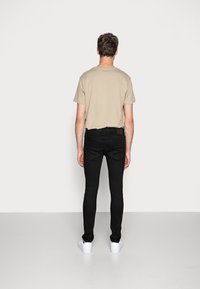 Mladý muž stojí čelem ke bílé zdi, oblečený v béžovém tričku, černých slim-fit džínech a bílých teniskách na bílé podlaze.