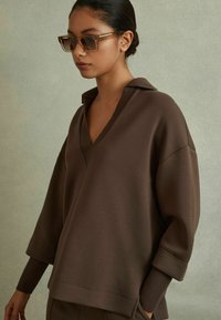 Maglione marrone con scollo a V e vestibilità oversize. Presenta maniche lunghe con polsini a coste, realizzato in un morbido tessuto testurizzato.