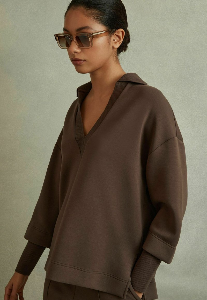 Maglione marrone con scollo a V e vestibilità oversize. Presenta maniche lunghe con polsini a coste, realizzato in un morbido tessuto testurizzato.