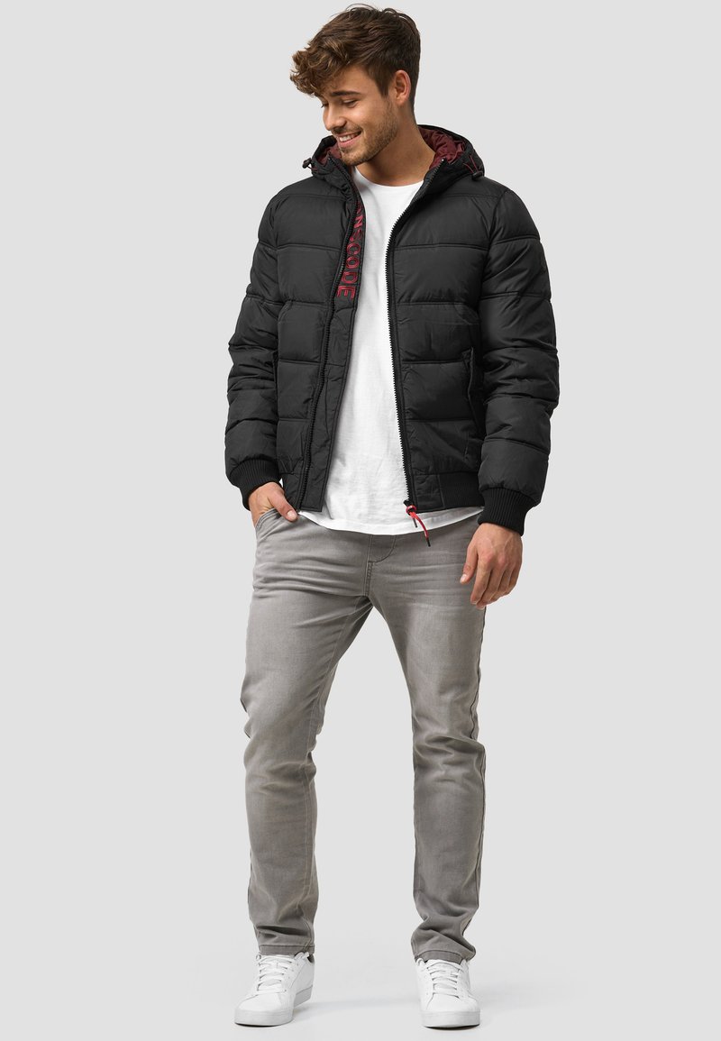 Schwarze gepolsterte Jacke mit einem Reißverschluss, ausgestattet mit einer bordeauxrot gefütterten Kapuze, gerippten Bündchen und einem bestickten Etikett. Kombiniert mit hellgrauen Jeans und weißen Sneakern.