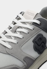 Baskets gris et blanches en tissu mesh, avec des accents en daim, des lacets plats blancs et un patch logo texturé en gris foncé sur le côté.