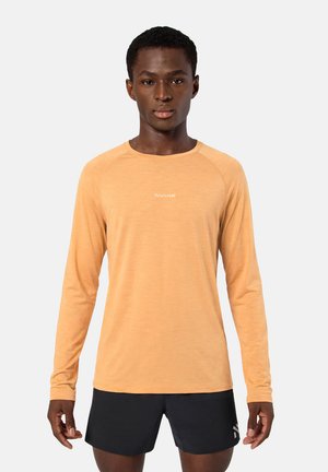 Joven con una camisa de manga larga color melocotón y pantalones cortos negros, de pie mirando hacia adelante sobre un fondo liso.