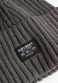 Bonnet gris tricoté à côtes avec une étiquette noire. L'étiquette comporte un texte blanc : "SUPERDRY UTILITY WEAR 360-366 / 054 MFG/CO." Texture douce.