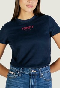 Tmavě modré bavlněné tričko s červeným logem "TOMMY JEANS", kulatým výstřihem, krátkými rukávy a přiléhavým střihem. Kombinováno s džínami.
