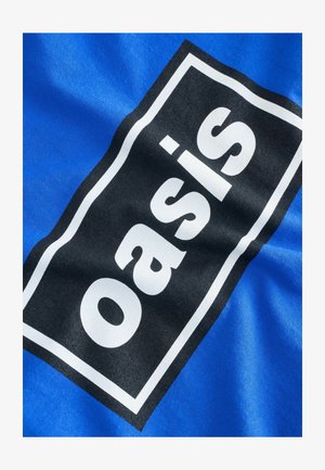 Zils audums ar spilgtu melnu taisnstūra logotipu, kurā redzams vārds "oasis" baltā krāsā, uz gludas tekstila faktūras.