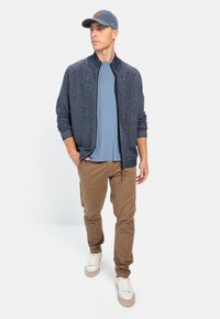 Navy-blauer Strickcardigan mit hohem Kragen, hellblauem T-Shirt, braunen Cargo-Hosen und weißen Sneakers, ergänzt durch eine blaue Kappe.