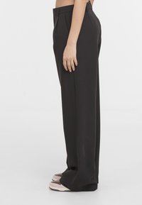 Vue latérale d'une personne portant un pantalon noir ample taille haute avec des plis, associé à des baskets beige et marron, sur fond blanc.