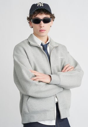 Felpa grigia con collo alto e zip frontale, indossata sopra una camicia bianca e cravatta blu; occhiali da sole scuri e un berretto a righe completano il look.
