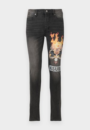 Mann trägt ein abgenutztes dunkelgraues T-Shirt mit dem Schriftzug „SIBI“, schwarze Jeans mit einem flammenden Totenkopf-Patch und schwarze, weiße und gelbe High-Top-Sneakers.