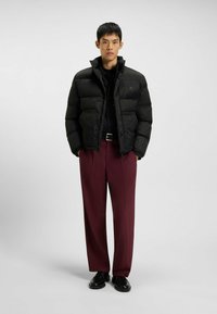 Giacca a vento nera con texture a motivi, colletto alto e zip frontale. Abbinata a pantaloni bordeaux e scarpe nere.