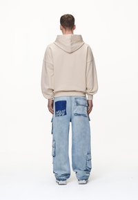 Hombre con sudadera beige y pantalones cargo sueltos de color azul claro, con el texto "Nunca es Demasiado Tarde" en un bolsillo trasero, de espaldas.