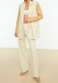 Beige Sakko mit hellbraunen Ärmeln, ausgestattet mit einem Doppelknopf-Design und zwei Fronttaschen. Dazu passende weit geschnittene Hosen.