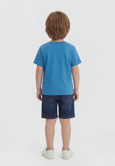Enfant aux cheveux châtain clair debout de dos, portant un t-shirt bleu à manches courtes, un short en jean foncé et des baskets blanches sur un fond uni.