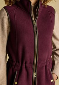 Burgunderfarbene Fleece-Weste mit hohem Kragen, Reißverschluss und braunen Akzenten. Verfügt über Seitentaschen mit goldenen Druckknöpfen. Weicher Stoff.