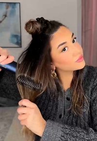 Black comb with wide teeth held in a hand, brushing long, straight hair. Person wears a dark knit sweater and has a top bun hairstyle.