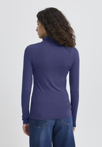 Donna con lunghi capelli ricci, vestita con un collo alto aderente blu scuro e jeans a gamba larga blu, in piedi di spalle contro uno sfondo neutro.