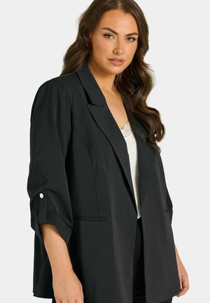 Donna con capelli lunghi e ondulati che indossa un blazer nero con maniche arrotolate sopra un top bianco con bordatura in pizzo su uno sfondo chiaro.