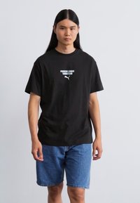 Sort bomulds t-shirt med hvid tekst "Porsche League Turbo Club" og Puma-logo, parret med blå denimshorts. Enkel, afslappet stil.