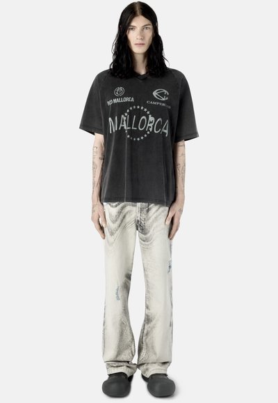 Personne aux longs cheveux noirs portant un t-shirt noir délavé Mallorca, un jean large clair et des chaussures noires sans lacets, debout devant un fond blanc.