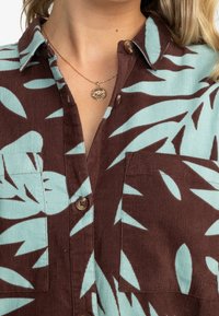 Chemise en velours côtelé brun avec un grand motif de feuilles vert menthe, fermetures à boutons et deux poches avant.