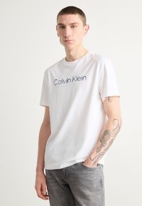 Balta kokvilnas T-kreklā ar "Calvin Klein" logotipu zilā krāsā, apaļā kakla izgriezums, īsas piedurknes, kopā ar gaiši pelēkiem džinsiem.