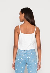 Top branco sem mangas com alças ajustáveis; o tecido possui uma textura suave. Usado com jeans de cintura alta azul claro com bordados florais.