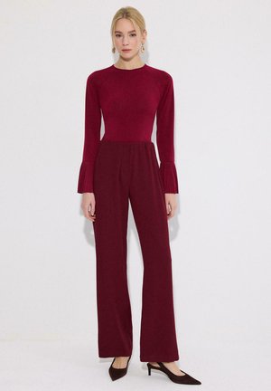 Žena s blond vlasmi, ktorá má oblečený priliehavý bordový dlhý rukáv s plisovanými manžetami a zodpovedajúce široké nohavice na bielom pozadí.