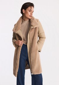 Beżowy płaszcz z mieszanych materiałów z pluszowym, teksturowanym futrzanym kołnierzem i wnętrzem, z zamkiem błyskawicznym i wyraźną sylwetką. Noszony na beżowej bluzce i niebieskich dżinsach.