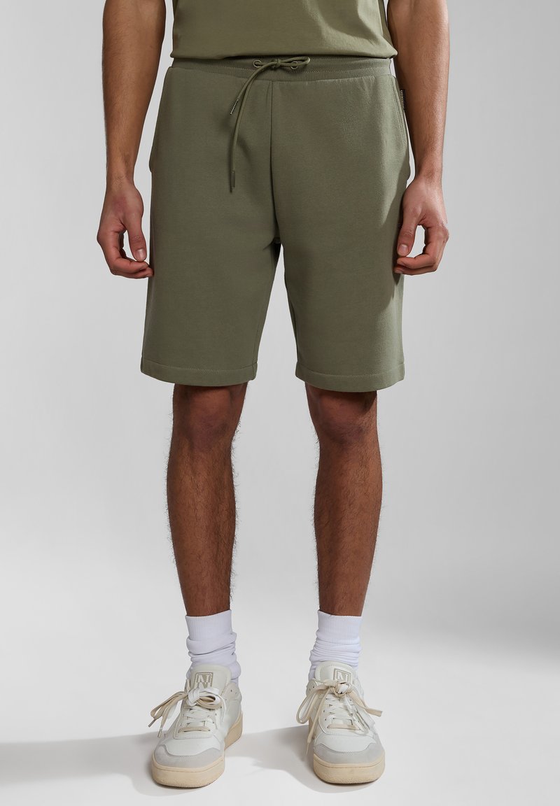 Pantalones cortos de algodón verde oliva con cintura ajustable, bolsillos laterales y dobladillos enrollados. Llevados con zapatillas blancas y calcetines blancos de caña baja.