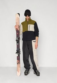 Uomo che indossa scarponi da sci neri, berretto nero, occhiali da sci scuri, giacca color block e pantaloni, che tiene uno sci in verticale contro uno sfondo bianco.