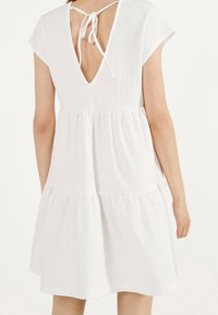 Vit kortärmad klänning med V-formad rygg och knytning, med en flerlagers kjol och strukturerad textil.