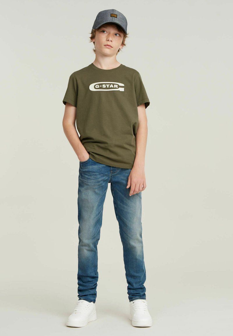 Olivegrøn bomuld t-shirt med hvid "G-STAR" logo, parret med lyseblå denim jeans og hvide sneakers. Personen har en grå kasket på.
