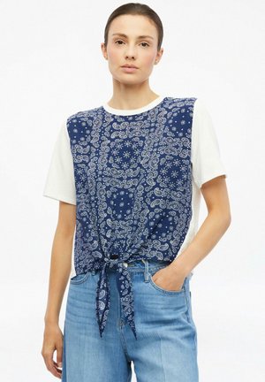 Junge Frau trägt ein weißes kurzärmliges Oberteil mit gebundener dunkelblauer Paisley-Weste und hellblauen Jeans, steht mit einer Hand in der Tasche.
